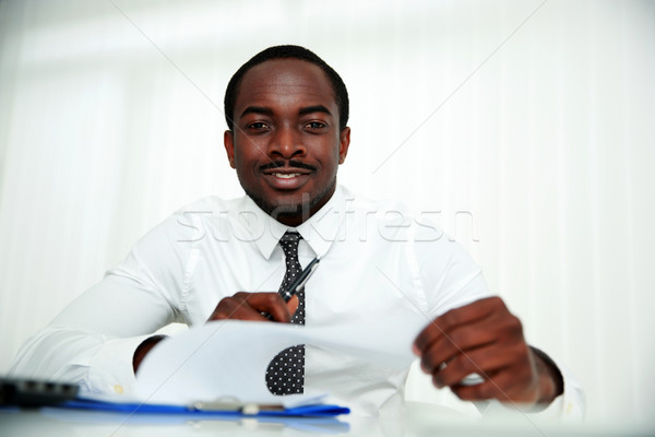 Happy african man sitting at the table and signing document in office Stock photo © deandrobot