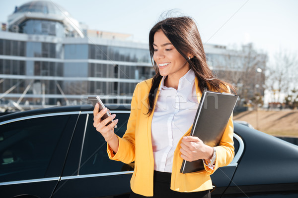 Smiling young businesswoman with documents in folder using smartphone outdoors Stock photo © deandrobot