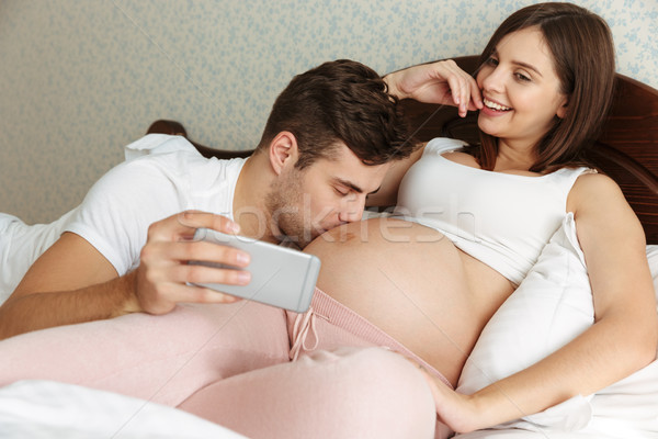 Attractive young pregnant couple taking selfie while lying in bed Stock photo © deandrobot