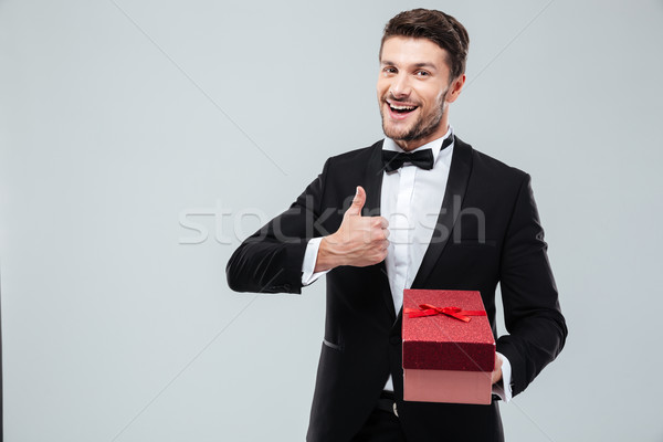 Stock photo: Man in tuxedo holding present box and showing thumbs up