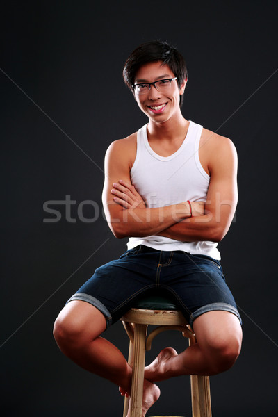 Happy asian man in glasses sitting on the chair over black background Stock photo © deandrobot