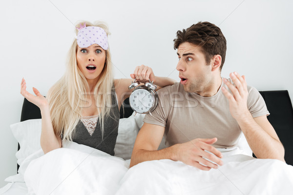 Shocked young lovers sitting in bed with opened mouth and holding alarm clock Stock photo © deandrobot