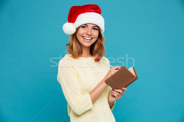 Close-up portrait of cheerful brunette woman in Santa's hat writing notes while looking at camera Stock photo © deandrobot