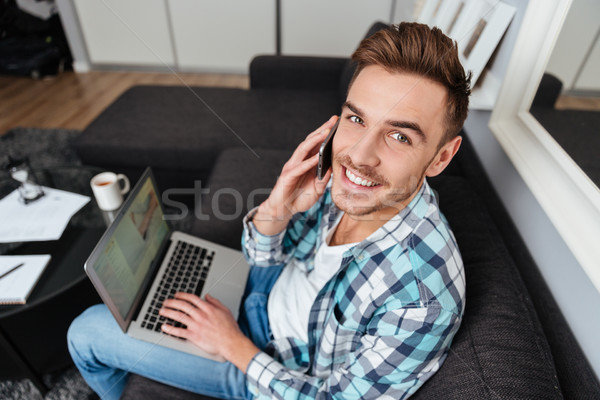 Cheerful man using laptop while talking by his phone. Stock photo © deandrobot