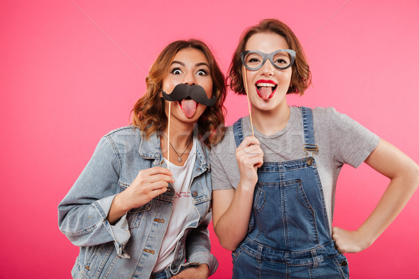 Funny ladies friends holding fake moustache and glasses. Stock photo © deandrobot
