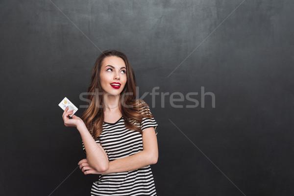 Thinking young lady standing over grey wall holding debit card Stock photo © deandrobot