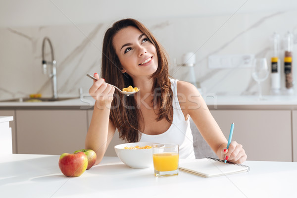Smiling thoughtful woman having breakfast cereal and writing in notepad Stock photo © deandrobot
