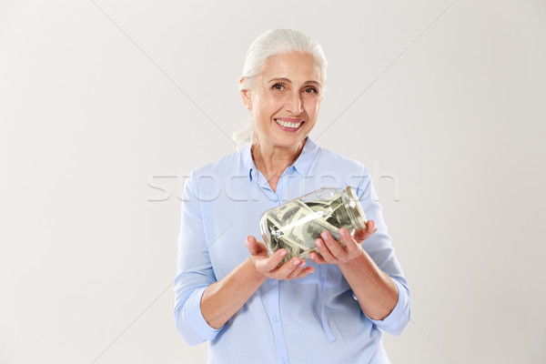 Happy senior lady in blue shirt holding glass bank with dollars, Stock photo © deandrobot