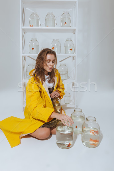 Lovely woman in raincoat sitting near jars with gold fishes Stock photo © deandrobot