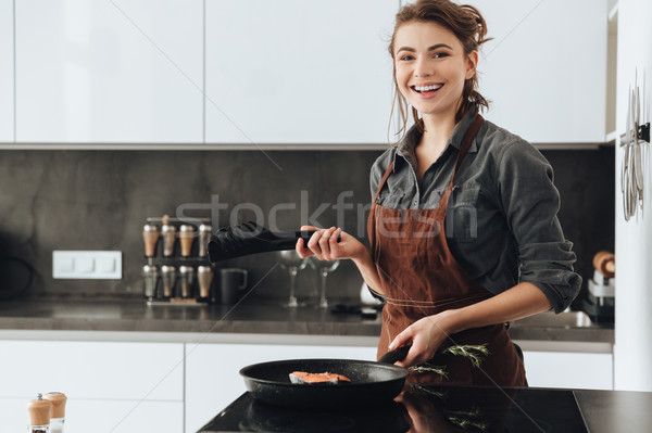 Stock photo: Happy lady standing in kitchen while cooking fish