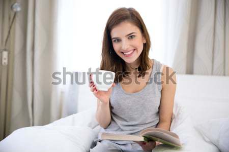 Stock photo: Smiling woman reading book and holding cup with coffee on the bed