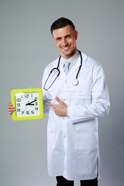 Portrait of a happy male doctor showing on clock on gray background Stock photo © deandrobot