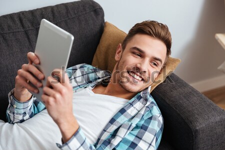 Cheerful bristle man lies on sofa and using tablet computer Stock photo © deandrobot