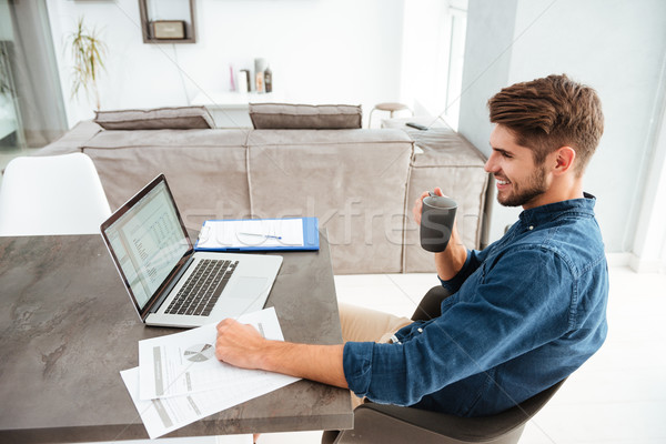 Cheerful young man drinking coffee while sitting near laptop Stock photo © deandrobot