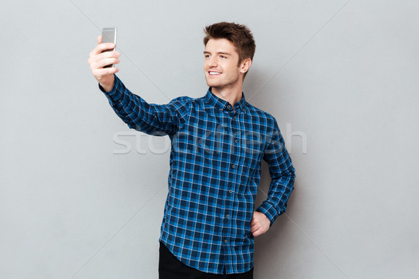 Cheerful young man standing over grey wall and make selfie Stock photo © deandrobot