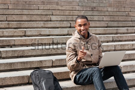Young african cheerful man sitting on staircase and using laptop Stock photo © deandrobot