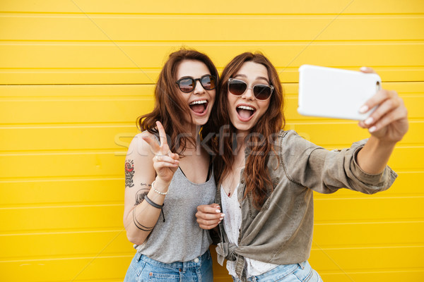 Stock photo: Young happy women friends standing over yellow wall make selfie