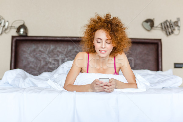 Cute lovely curly woman using mobile phone lying in bed  Stock photo © deandrobot