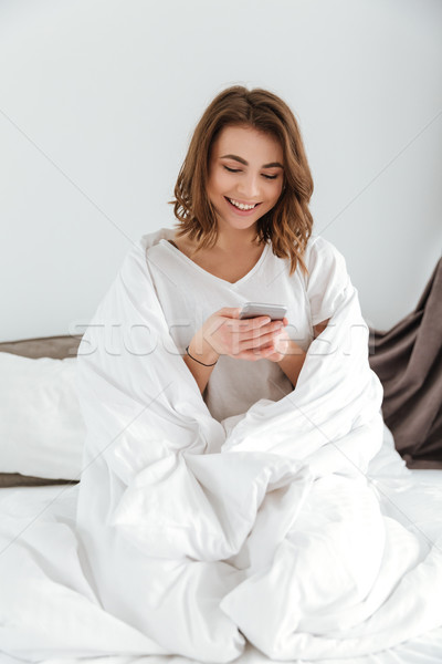 Happy beautiful young woman sitting and using mobile phone in bed Stock photo © deandrobot