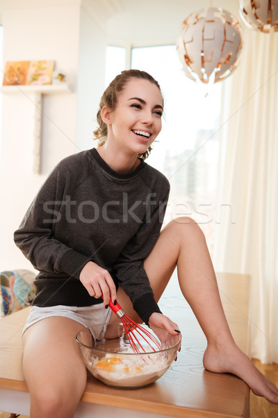 Young laughing woman sitting on kitchen table and whisking eggs Stock photo © deandrobot