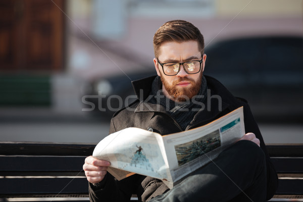 Concentrated young man sitting on bench and reading newspaper outdoors Stock photo © deandrobot