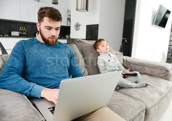 Serious bearded father using laptop while his son watching TV. Stock photo © deandrobot