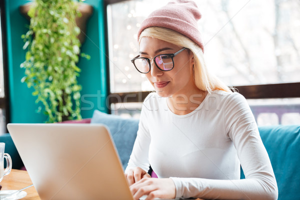 Cheerful young lady wearing hat using laptop in cafe. Stock photo © deandrobot