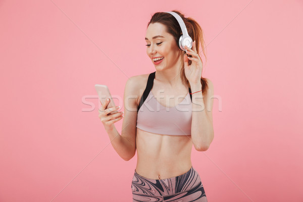 Stock photo: Pleased sportswoman in headphones listening music and using smartphone