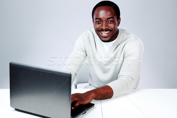 Laughing african man sitting at the table and using laptop on gray background Stock photo © deandrobot