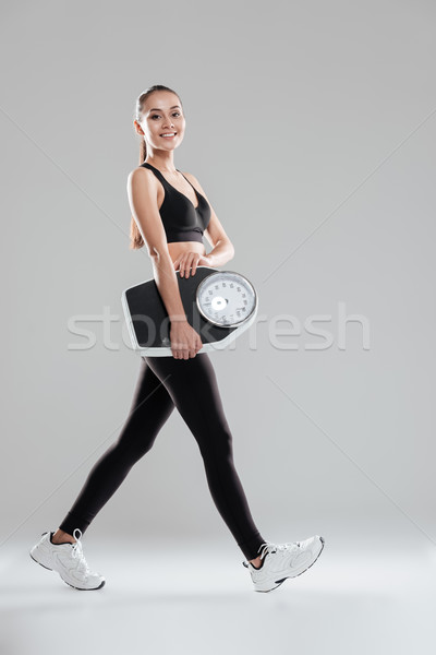 Happy cute young woman athlete walking and holding weighing scale Stock photo © deandrobot