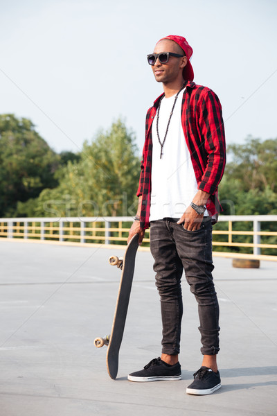 Young dark skinned man wearing sunglasses holding the skateboard Stock photo © deandrobot