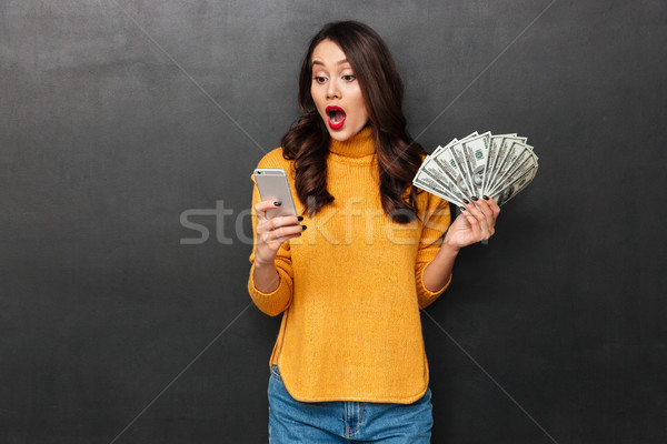 Surprised brunette woman in sweater holding money and using smartphone Stock photo © deandrobot