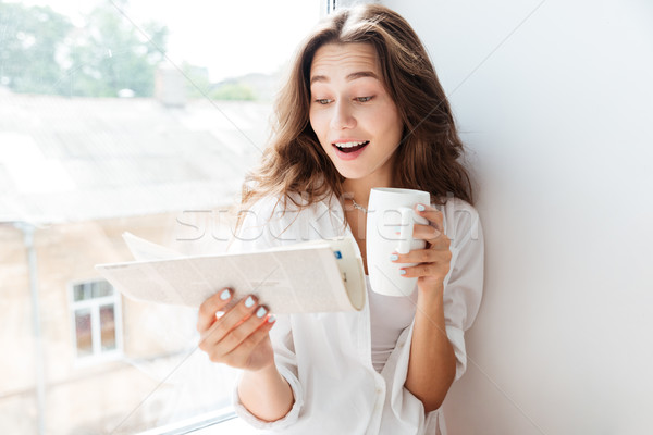 Happy young bruette woman looking at newspaper and drinking tea Stock photo © deandrobot