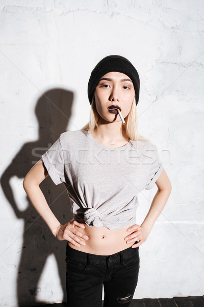 Stylish young woman hipster in black hat standing and smoking Stock photo © deandrobot