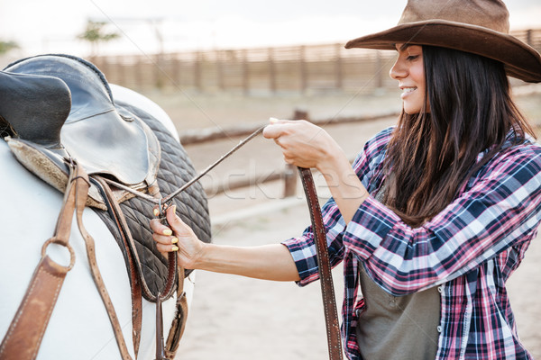 Woman cowgirl standing and putting saddle on horse Stock photo © deandrobot