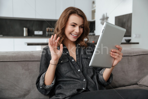 Happy lady sitting on sofa looking at tablet and waving. Stock photo © deandrobot