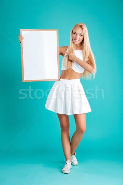 Smiling blonde woman standing and pointing finger at blank board Stock photo © deandrobot