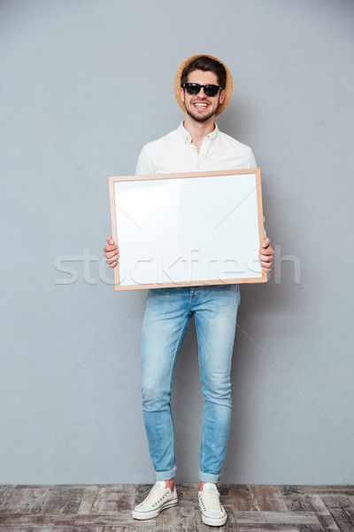 Smiling young man in hat and sunglasses holding white board Stock photo © deandrobot