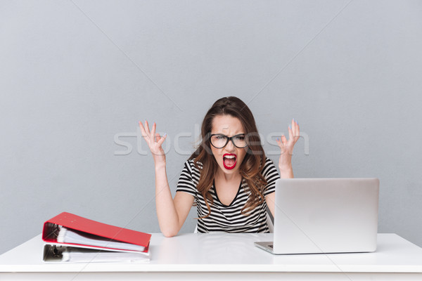 Angry screaming young lady wearing glasses sitting over grey wall. Stock photo © deandrobot
