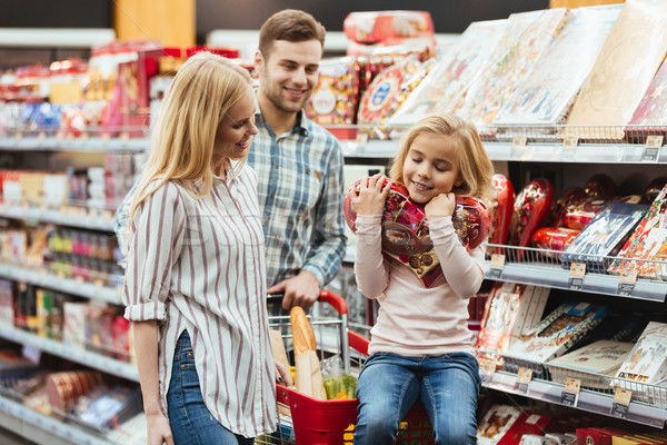 Smiling little girl sitting on a shopping cart and choosing candy with her parents at the supermarke Stock photo © deandrobot