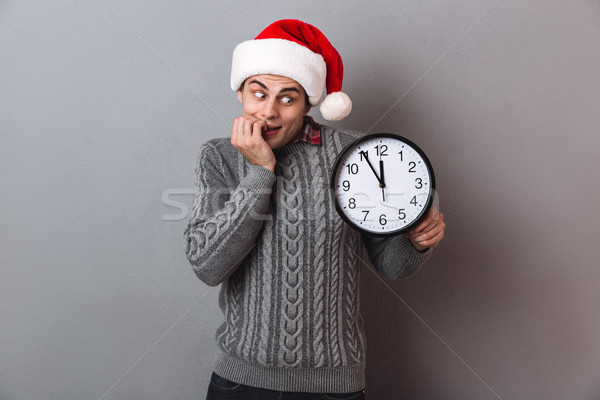 Stock photo: Intrigued man in sweater and christmas hat holding clock