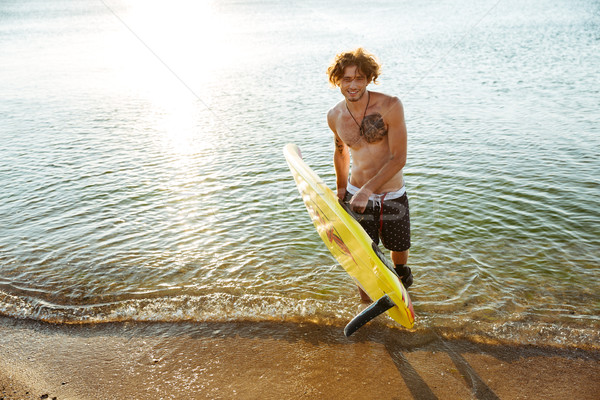 Smiling curly man holding his surf board at the beach Stock photo © deandrobot