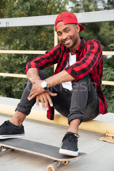 Cheerful dark skinned guy wearing cap sitting with his skateboard Stock photo © deandrobot