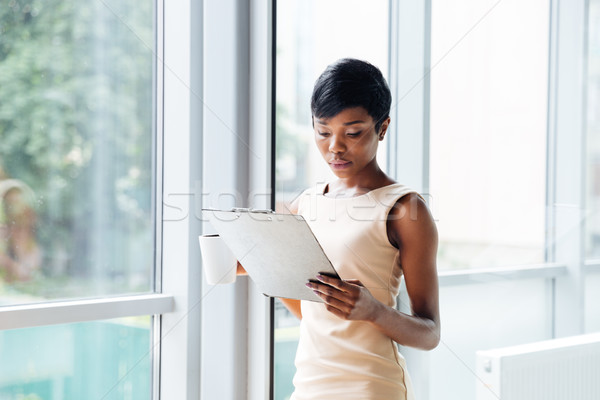 Beautiful african young businesswoman holding clipboard and reading in office Stock photo © deandrobot