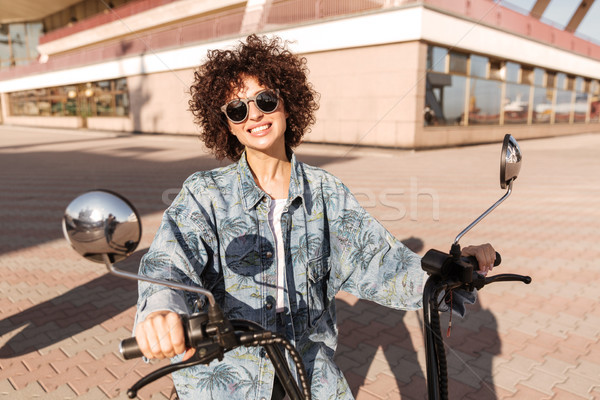Image of joyful curly woman in sunglasses sitting on motorbike Stock photo © deandrobot