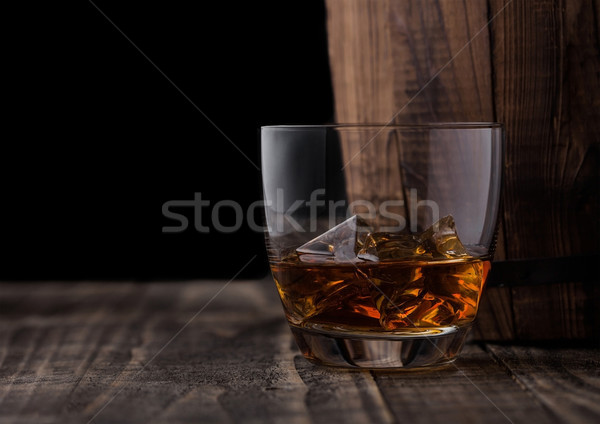 Glass of whiskey with ice cubes next to wooden barrel. Cognac brandy drink Stock photo © DenisMArt