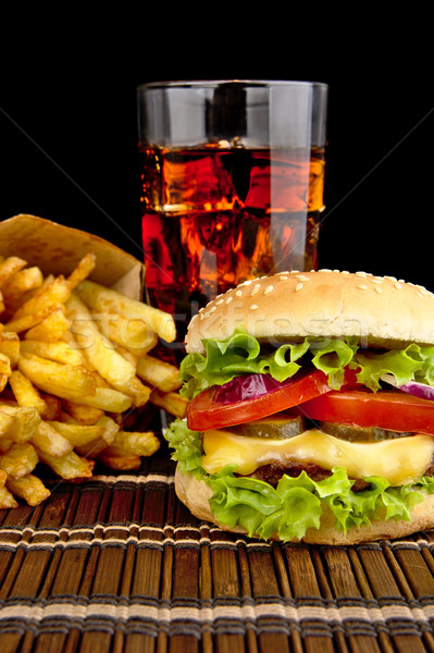 Stock photo: Big cheeseburger with french fries with glass of cola on wooden mat on black