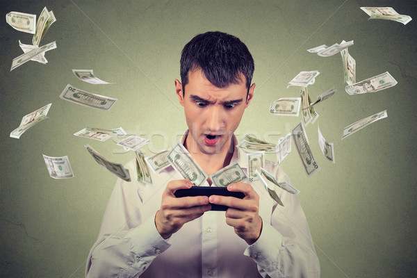 Shocked young man using smartphone with dollar bills banknotes flying away Stock photo © ichiosea