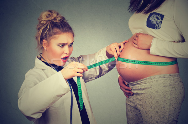 Stock photo: Pregnant woman exposing belly while a shocked gynecologist doctor uses measuring tape to follow grow