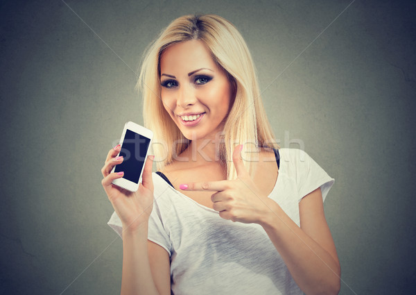 Smiling young woman is pointing at smartphone standing on gray background. Stock photo © ichiosea